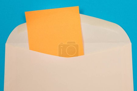 Foto de Sobre beige con hoja de papel naranja en blanco en el interior, acostado sobre fondo azul: finge con espacio de copia. Recibir correo o aviso, enviar postal - Vista superior, disposición plana - Imagen libre de derechos