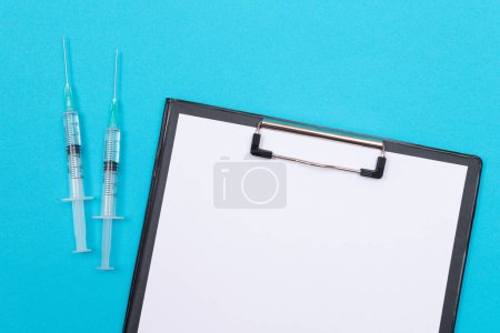 Téléchargez les photos : Concept de vaccination, d'immunologie ou de révocation : deux seringues médicales posées sur la table bleue dans un cabinet médical d'un hôpital ou d'une clinique. Presse-papiers noir avec feuille de papier - Maquette avec espace de copie - en image libre de droit