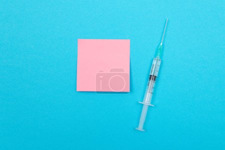 Téléchargez les photos : Concept de vaccination, d'immunologie ou de revaccination - Une seringue médicale jetable posée sur la table bleue dans un cabinet médical d'un hôpital ou d'une clinique. Note collante rose vierge - Maquette avec espace de copie - en image libre de droit
