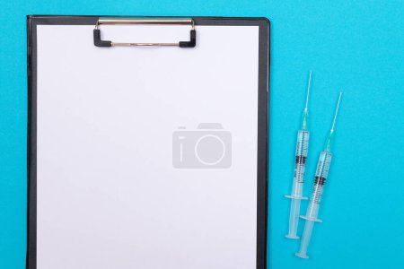 Téléchargez les photos : Concept de vaccination, d'immunologie ou de révocation : deux seringues médicales posées sur la table bleue dans un cabinet médical d'un hôpital ou d'une clinique. Presse-papiers noir avec feuille de papier - Maquette avec espace de copie - en image libre de droit