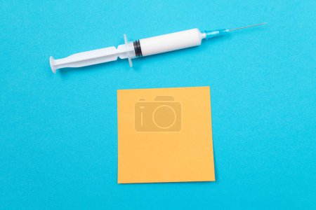 Téléchargez les photos : Concept de vaccination, d'immunologie ou de revaccination - Une seringue médicale jetable posée sur la table bleue dans un cabinet médical d'un hôpital ou d'une clinique. Note collante orange vierge - Maquette avec espace de copie - en image libre de droit