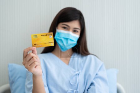 Foto de Mujer asiática feliz con una máscara médica y la celebración de la maqueta de tarjeta de crédito / tarjeta de seguro y signo ok en una cama de hospital. Póliza de seguro por banco, concepto de tratamiento médico de pago - Imagen libre de derechos