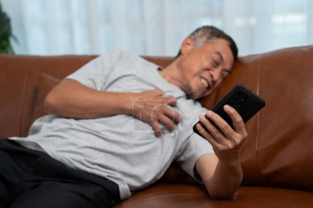 Senior mâle asiatique presse la main à sa poitrine a une crise cardiaque souffre de douleurs insupportables dans le salon, les hommes seuls essaient d'utiliser le smartphone pour appeler à l'aide d'urgence.