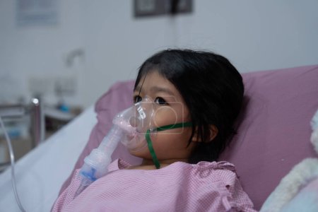 A little Asian girl has an oxygen mask and breathing through a nebulizer at the hospital. Concept of bronchitis, respiratory and Medical treatment, inhaling medicine, mist, from a nebulizer.