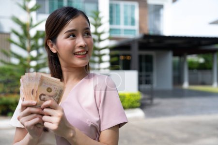Portrait of happy attractive Asian women holding a lot of banknotes in front of home to celebrate and Pay the final installment of the house, Loans for real estate concept, Smiling young successful