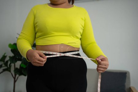 Foto de Mujer con sobrepeso usando una cinta métrica alrededor de la cintura y la grasa del vientre, entrenamiento de la mujer asiática gorda en casa. Concepto de saludable, comida para la salud y el estilo de vida, pérdida de peso - Imagen libre de derechos