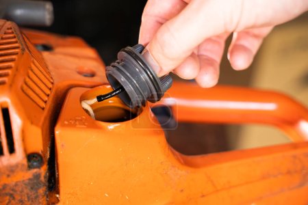 A hand unscrews the cap of the fuel mixture tank in an orange chainsaw. Gas tank for gasoline of a two-stroke engine close-up. Refueling a chainsaw