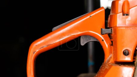Orangefarbener Kettensägengriff mit Bedienelementen in Großaufnahme auf dunklem Hintergrund. Die Taste, um den Motor mit Kraftstoff zu versorgen und die Drehzahl bei einem Zweitaktmotor zu erhöhen. Altes Schneidwerkzeug
