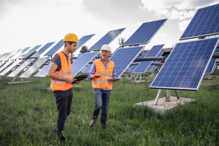 portrait of two adult male engineers wearing safety vest consulting with blue print in front of solar panels. worker in solar plant area, renewable and green energy concept. Portrait