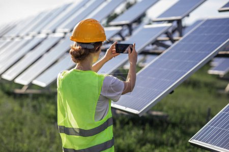 Foto de Attractive woman ecological engineer with safety equipment at photovoltaic solar farm taking pictures of solar panels. - Imagen libre de derechos