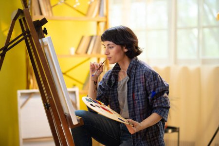 Téléchargez les photos : Une peintre femme est très concentrée et semble heureuse d'elle-même car elle travaille à peindre quelque chose de très agréable sur une toile. - en image libre de droit