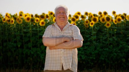 Téléchargez les photos : Un vieil homme en chemise à carreaux se tient debout les bras croisés dans un beau champ de tournesol et sourit joyeusement. - en image libre de droit