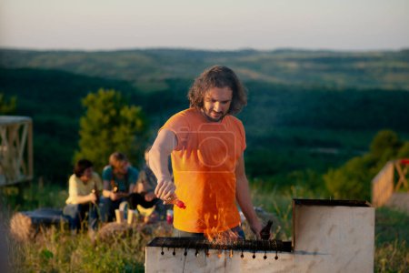 Foto de A very handsome man is cooking something at an outdoor grill while a few people behind him are sharing drinks outdoors in the nature. - Imagen libre de derechos
