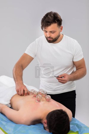 Photo for Professional therapist man get his vacuum cups and put them over the back of his client while the man are laying down on the massage table. Portrait - Royalty Free Image