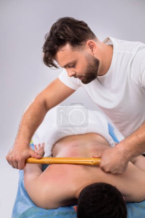 Téléchargez les photos : Prenant la forme vidéo du haut au salon de spa l'homme thérapeute en utilisant le bâton en bois pour bien masser les muscles du dos et obtenir de bons résultats pour son client. Portrait - en image libre de droit