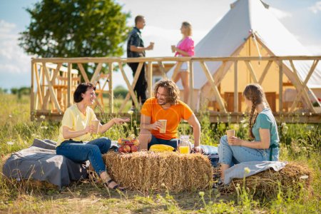 Photo for Campsite very charismatic and attractive ladies and guys have fun time together they drinking and looking over the smartphone watching something together while sitting down on the haystack. Portrait - Royalty Free Image