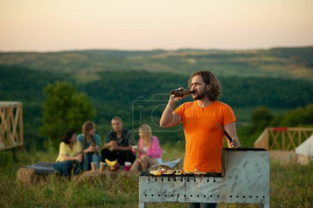 Photo for A handsome middle aged man is flipping over vegetables as he is cooking them on a grill outside in the nature with his friends that are sitting behind him. Portrait - Royalty Free Image