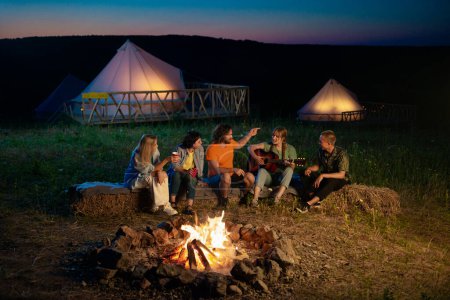 Téléchargez les photos : Au camping groupe d'amis très excitants et beaux multiraciaux assis sur la meule de foin chanter et jouer à la guitare boire de la bière et danser. Tente - en image libre de droit