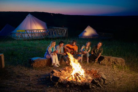 Foto de Muy bonita dama tocando en la guitarra con sus amigos en el camping junto a la hoguera mientras se sienta en el pajar. - Imagen libre de derechos