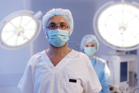 Téléchargez les photos : A handsome male surgeon with glasses is in a medical hospital room with a nurse standing behind him in very good lighting. - en image libre de droit
