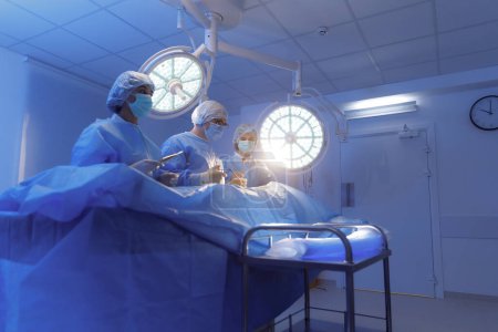 Foto de Un cirujano masculino está usando una pinza para operar a su paciente mientras está en una sala de cirugía y recibe ayuda de sus dos asistentes.. - Imagen libre de derechos