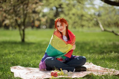 Téléchargez les photos : Belle lesbienne tenant le drapeau arc-en-ciel lgbt assis sur l'herbe au milieu du parc et profitant du temps avec elle-même. - en image libre de droit