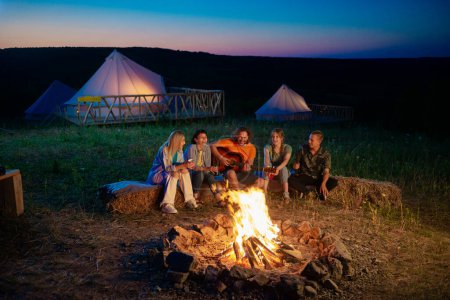 Téléchargez les photos : Groupe d'amis charismatiques et attrayants se réunissant au camping le soir ils font un feu de camp prendre un siège sur la meule de foin et jouer à la guitare et chanter tous ensemble. Tente - en image libre de droit