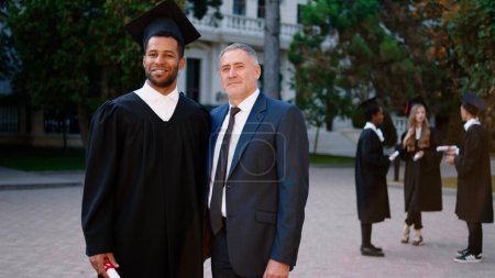 Photo for Multiethnic concept in the college garden posing in front of the camera mature man college professor and his graduate student with the diploma. - Royalty Free Image