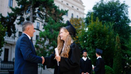 Foto de Muy emocionada rubia se gradúa con gorra de graduación y traje tomando su diploma del profesor universitario en el día de la graduación y luego posando frente a la cámara con una gran sonrisa. ARRI - Imagen libre de derechos