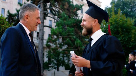 Foto de Hombre apuesto graduado tener una conversación con su padre después de la graduación que sostiene su diploma que discuten en el jardín de la universidad. - Imagen libre de derechos