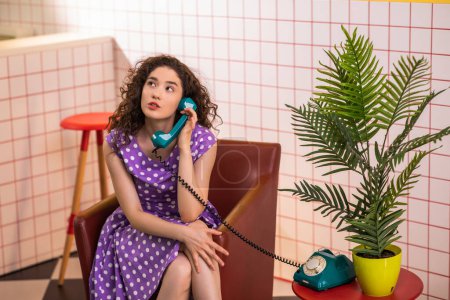 Photo for Pretty young lady in a pink dress sitting on the sofa in the retro cafe and discussing with someone on the vintage phone. Retro - Royalty Free Image