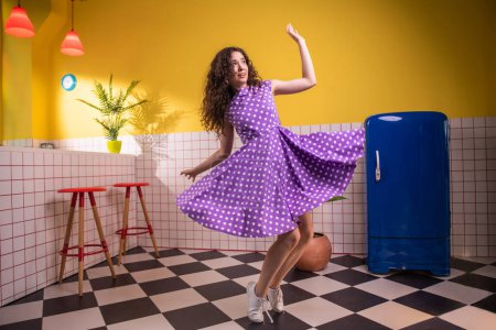 Téléchargez les photos : Femme charismatique incroyable avec un grand sourire se déplaçant très excité et dansant devant la caméra dans la cuisine, elle porte une belle robe rose. Rétro - en image libre de droit