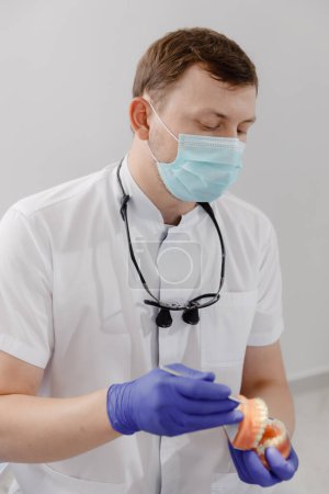 Photo for Closeup to the camera good looking dentist with protective mask explains the process of fixing the braces to teeth he using the tooth model for this. Dentist - Royalty Free Image
