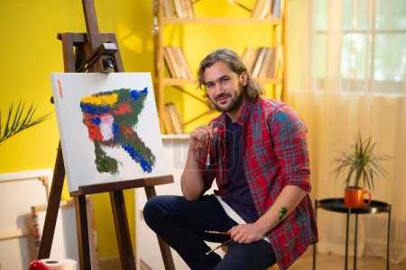 Foto de Un pintor muy guapo está parado frente a su pintura y sonriendo a la cámara. pintor - Imagen libre de derechos