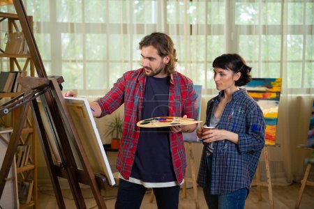 Photo for In the art studio charismatic and beautiful couple painting a picture on canvas together using the pallet and add some oil colour on the canvas. - Royalty Free Image