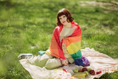 Photo for In a sunny day beautiful young woman in the park laying down on the blanket down on the grass and smiling pretty to the camera she holding the lgbt flag. Lgtb - Royalty Free Image