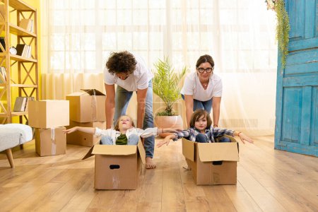 Photo for In the new home happy and excited parents take their two little kids and put them on the boxes and start to make a ride through the living room . Family - Royalty Free Image
