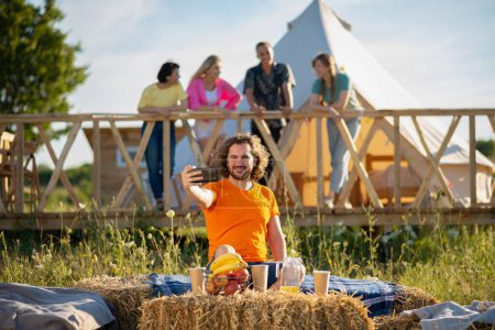 Photo for At the campsite great looking man looking over his smartphone while sitting down on the haystack he get excited while his friends chilling on the background. Friends - Royalty Free Image