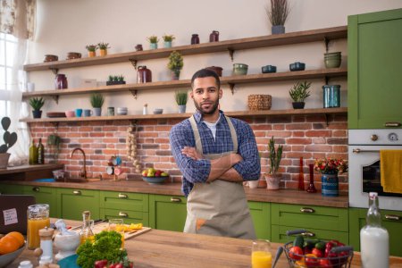 Photo for In front of the camera posing guy at the kitchen he looking straight and smiling very charismatic he wear kitchen apron and crossing hands. - Royalty Free Image