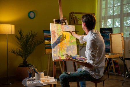 Foto de En el estudio de arte artista hombre guapo tomar el pincel de pintura y empezar a pintar nueva imagen en el lienzo mientras está sentado en la silla. Pintura - Imagen libre de derechos