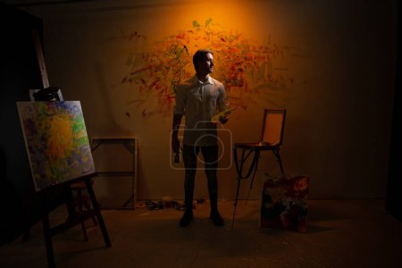 Foto de Delante de la cámara captura el retrato de un artista posando y mirando recto mientras sostiene su paleta de colores al óleo. - Imagen libre de derechos
