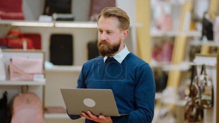 Téléchargez les photos : Superviseur homme charismatique dans la boutique d'accessoires en utilisant un ordinateur portable numérique pour analyser la statistique des ventes de la boutique en ligne. - en image libre de droit