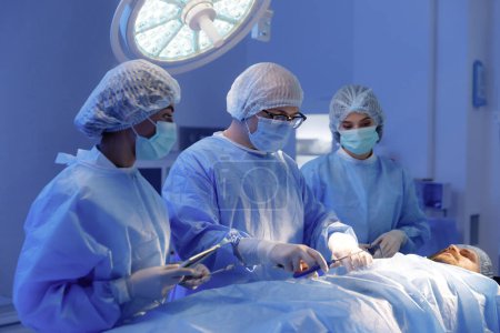 Téléchargez les photos : A surgeon and his two assistants are wearing surgical gear and operating on a patient. - en image libre de droit