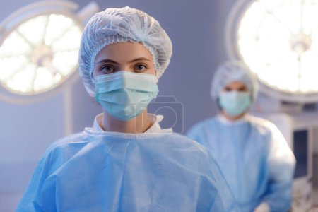Photo for A close up shot of a female nurse looking directly at the camera and looking beautiful, while there is medical head lights and a nurse standing behind her. - Royalty Free Image