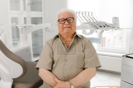 Foto de Paciente anciano carismático tiene un tratamiento de cuidado bucal en la clínica dental moderna posando frente a la cámara en el consultorio del dentista cruzando las manos y mirando directamente. Dentista - Imagen libre de derechos
