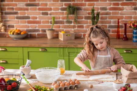 Téléchargez les photos : Devant la caméra petite fille charismatique à l'îlot de cuisine prendre la pâte et en utilisant le rouleau préparant son dessert préféré. Portrait - en image libre de droit