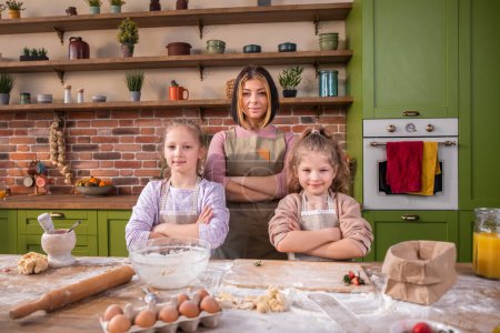Photo for Posing in front of the camera amazing looking woman and her two kids girls at the kitchen island they crossing hands and looking straight and smiling large. Portrait - Royalty Free Image