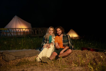 Téléchargez les photos : Closeup to the camera amazing looking couple have a romantic time at the campsite they sitting down on the haystack beside the fire stake and melt some marshmallows and start to eating, Portrait - en image libre de droit