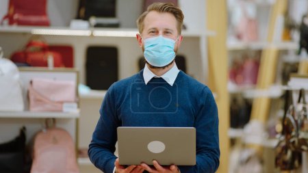 Téléchargez les photos : Vendeur charismatique dans la boutique d'accessoires portant un masque de protection et travaillant sur son ordinateur portable devant la caméra dans la pandémie de coronavirus. - en image libre de droit