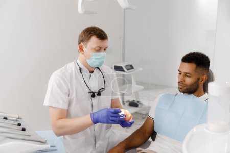 Foto de Un odontólogo guapo con máscara protectora explica al paciente el proceso de trabajo que utiliza un modelo de diente para mostrar y explicar más fácilmente. - Imagen libre de derechos